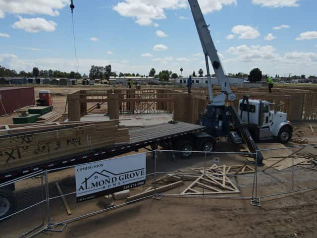 Aerial view of a crane truck and flatbed behind a temporary fence with an Almond Grove Homes sign, and a sheathed house behind