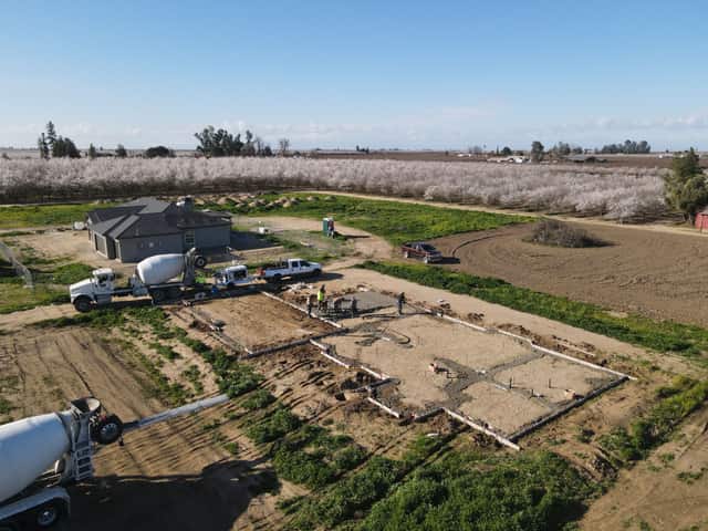Aerial view of a foundation pour with a cement mixer truck, a finished house, and a blooming almond orchard behind