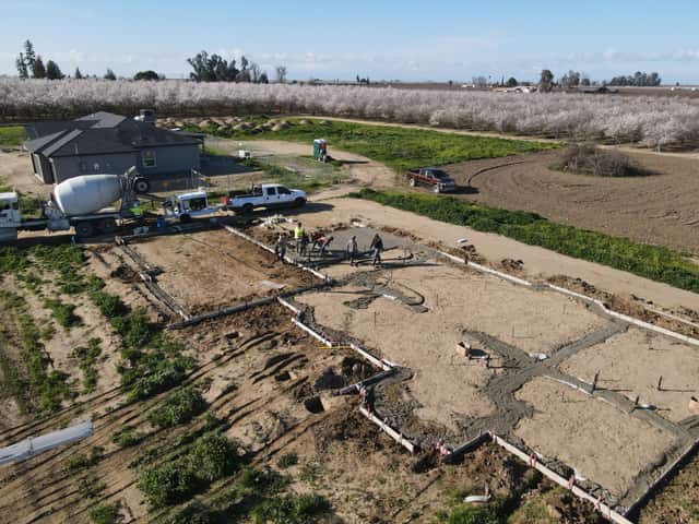 Aerial view of poured stem walls and graded building pad with crews at work and an almond orchard behind