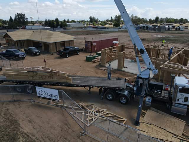 A flatbed with a stack of roof trusses and a crane beside a framed home, with a second sheathed house in the background