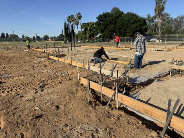 Crew members setting and checking forms on a footing at the edge of a foundation, with palm trees behind a fence