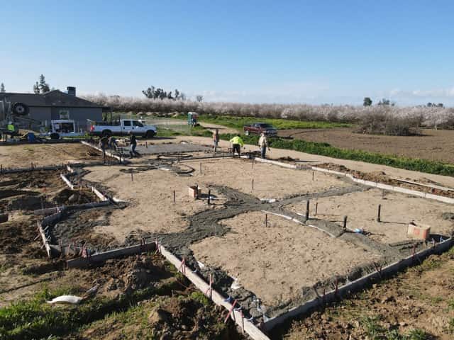 Low aerial view of a crew finishing a concrete foundation on a graded pad beside a blooming almond orchard