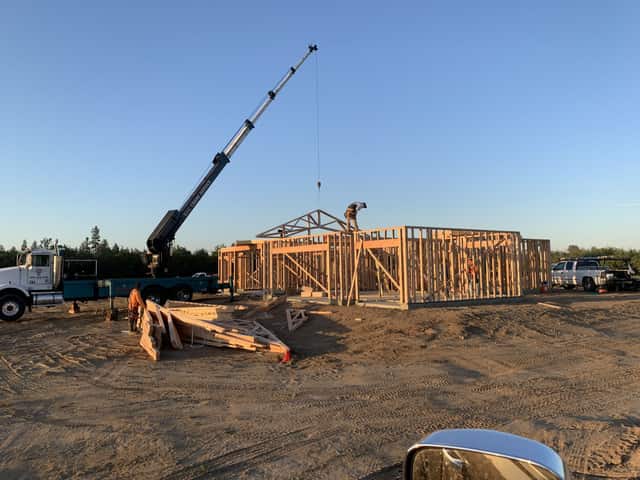Crane truck lifting trusses beside a framed single story home on a graded lot under a clear sky