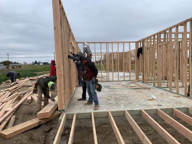 Framers lifting a wall section upright on the floor platform, with stacked joists in the foreground