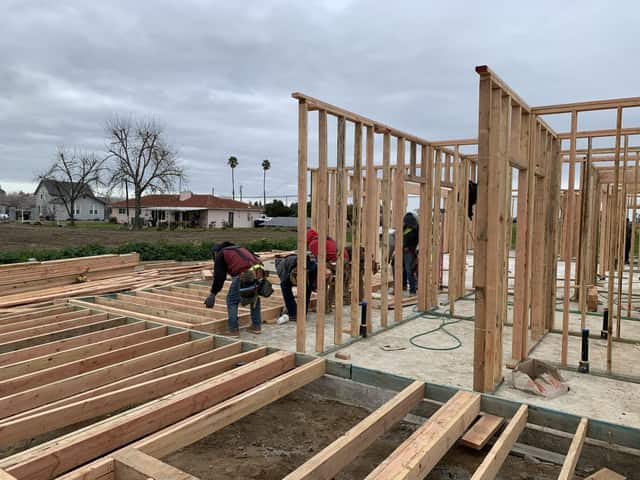 Framing crew setting wall sections on a platform floor of joists with a house and trees in the distance