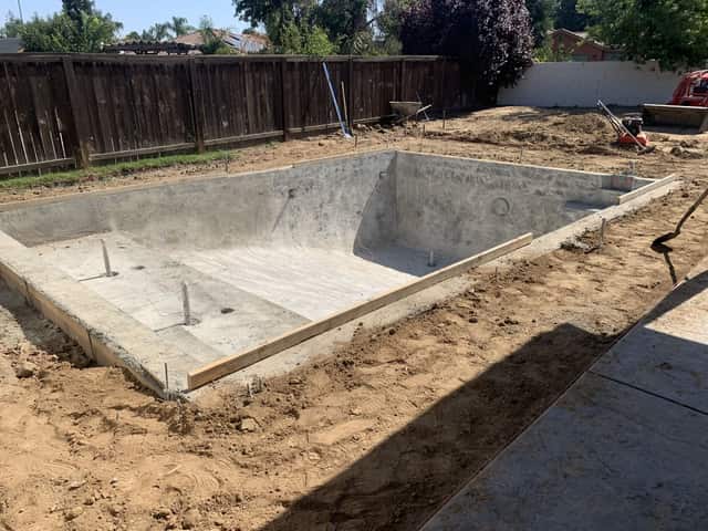A concrete pool shell with a bench and steps in a fenced backyard